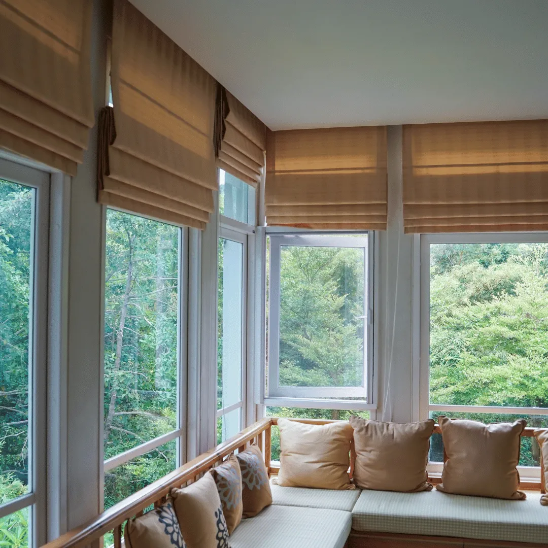 Tan folded Roman shades across a wraparound window seat overlooking trees